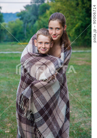 Young sister and brother with freckles on their faces stand covered with plaid in park, get warm, smiling and looking at camera. Young sister and brother with freckles on their faces stand covered with plaid in park, get warm, smiling and looking at camera. 90906714