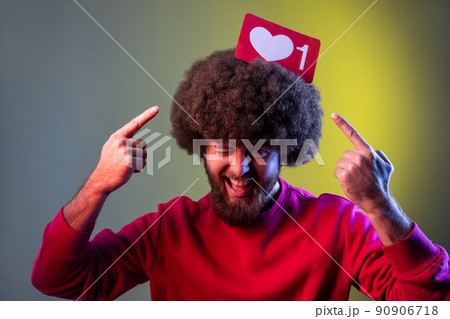 Hipster man holding pointing at Heart Like icon of social media on his head, notification button to follow, subscribe blog. Indoor studio shot isolated on colorful neon light background. Hipster man holding pointing at Heart Like icon of social media on his head, notification button to follow, subscribe blog. Indoor studio shot isolated on colorful neon light background. 90906718
