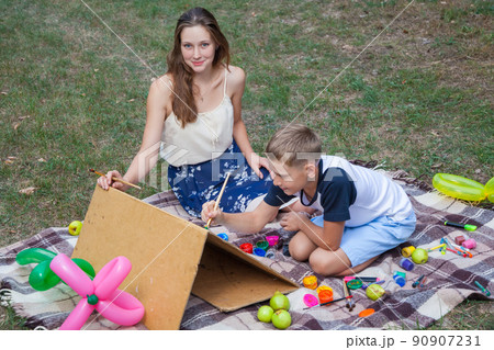Older sister trying to teach her brother to paint and posing in the park on summer time, looking at camera and smiling. 90907231