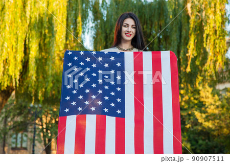 Beautiful young woman with classic dress holding american flag in the park. fashion model holding us smiling and looking at camera. usa lifestyle. 90907511