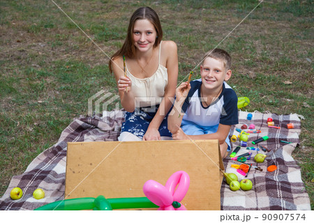 Older sister trying to teach her brother to paint and posing in the park on summer time, looking at camera and smiling. 90907574