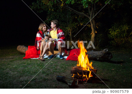 Young happy loving couple sitting on log near the fire covered with plaid holding wine, drinking and enjoy their romantic night. looking at each other and smiling. 90907579