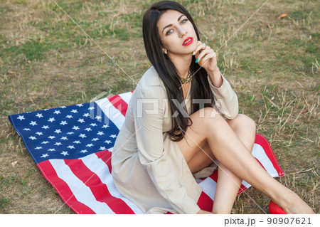 Beautiful sexy young woman with classic dress lying down on american flag in the park. fashion model holding us smiling and looking at camera. USA lifestyle with toothy smile. Beautiful sexy young woman with classic dress lying down on american flag in the park. fashion model holding us smiling and looking at camera. USA lifestyle with toothy smile. 90907621