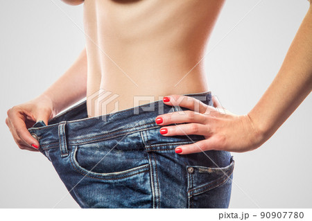 Closeup of slim waist of young woman standing in big jeans showing successful weight loss, indoor studio shot, isolated on light gray background, diet concept. 90907780