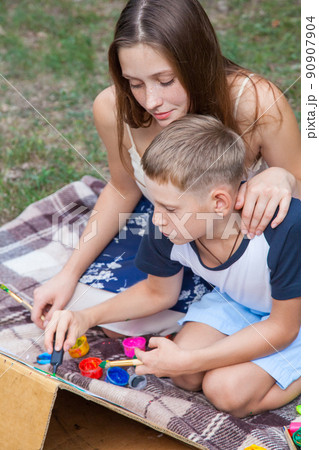 Older sister trying to teach her brother to paint and posing in the park on summer time, drawing and painting.. 90907904