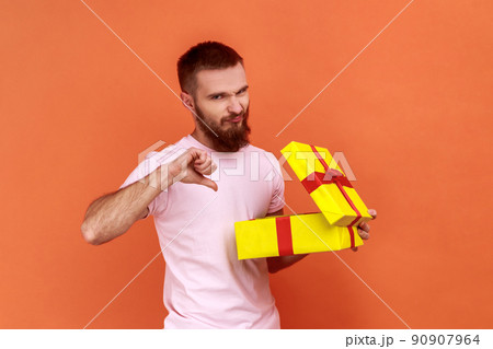 Portrait of bearded man unpacking present to know what he received, being frustrated and disappointed, showing thumb down, wearing pink T-shirt. Indoor studio shot isolated on orange background 90907964