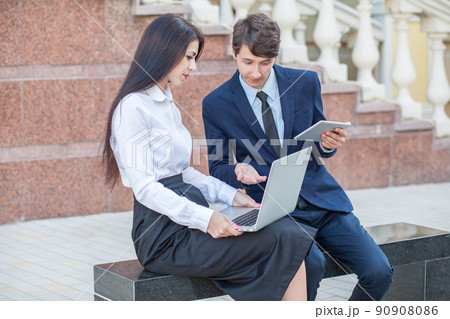 Boss in blue suit and his assistant in white blouse and black skirt discussing their work outdoors. 90908086