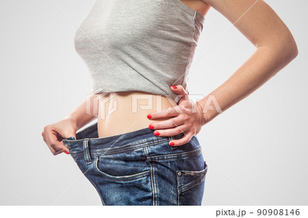Closeup of slim waist of young woman standing in big jeans and gray top showing successful weight loss, indoor studio shot, isolated on light gray background, diet concept. 90908146
