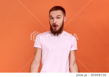 Portrait of amazed bearded man standing with mouth open in surprise, has shocked expression, hears unbelievable news, wearing pink T-shirt. Indoor studio shot isolated on orange background. Portrait of amazed bearded man standing with mouth open in surprise, has shocked expression, hears unbelievable news, wearing pink T-shirt. Indoor studio shot isolated on orange background. 90908156