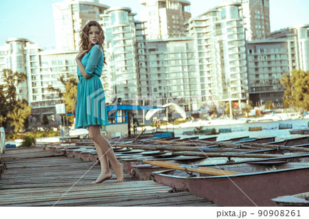 Beautiful young fashion model in green turquois dress and fashion makeup and hairstyle is posing on pier, posing and looking at camera on city background. Beautiful young fashion model in green turquois dress and fashion makeup and hairstyle is posing on pier, posing and looking at camera on city background. 90908261