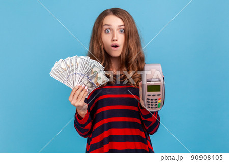 Astonished woman wearing striped casual style sweater, standing with shocked expression, showing pos payment terminal and dollars bills. Indoor studio shot isolated on blue background. Astonished woman wearing striped casual style sweater, standing with shocked expression, showing pos payment terminal and dollars bills. Indoor studio shot isolated on blue background. 90908405