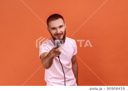 Portrait of bearded man posing with microphone in hands, offers mic, journalist asking questions, looking at camera, wearing pink T-shirt. Indoor studio shot isolated on orange background. Portrait of bearded man posing with microphone in hands, offers mic, journalist asking questions, looking at camera, wearing pink T-shirt. Indoor studio shot isolated on orange background. 90908459