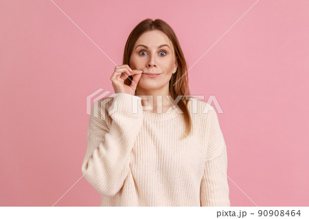 I won't tell anyone. Intimidated blond woman zipping lips and looking at camera, covering mouth promising to keep terrible secret, wearing white sweater. Indoor studio shot isolated on pink background 90908464