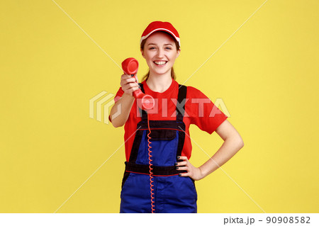 Portrait of affirmative worker woman holding out handset, asking call her and order service, looking friendly at camera, wearing overalls and red cap. Indoor studio shot isolated on yellow background. 90908582