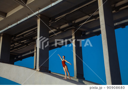 Beautiful young strong healthy woman ballerina gymnast in red leotard posing as model on bridge girder and blue sky and looking into the distance. 90908680