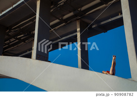 Beautiful young strong healthy woman ballerina gymnast in red leotard posing as model on bridge girder and blue sky and looking into the distance. 90908782