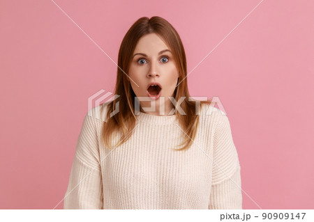 Portrait of shocked blond woman standing looking at camera with open mouth, being astonished by breaking news, wearing white sweater. Indoor studio shot isolated on pink background. Portrait of shocked blond woman standing looking at camera with open mouth, being astonished by breaking news, wearing white sweater. Indoor studio shot isolated on pink background. 90909147