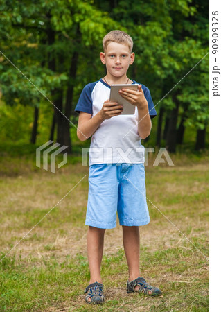 Cute young caucasian kid with freckles on his face in blue shorts and white and blue T-shirt and tablet and working in park and smiling. standing and looking at camera. 90909328