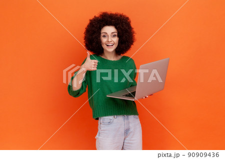Smiling woman with Afro hairstyle wearing green casual style sweater working on laptop, looking at camera and showing thumb up, positive emotions. Indoor studio shot isolated on orange background. 90909436