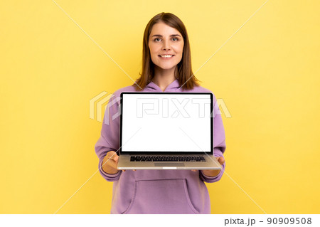 Positive optimistic woman standing showing laptop with white empty screen for promotion, looking at camera with smile, wearing purple hoodie. Indoor studio shot isolated on yellow background. 90909508