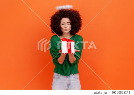 Positive funny angelic woman with Afro hairstyle wearing green casual style sweater giving wrapped present box, making kiss gesture, keeps eyes close. Indoor studio shot isolated on orange background. Positive funny angelic woman with Afro hairstyle wearing green casual style sweater giving wrapped present box, making kiss gesture, keeps eyes close. Indoor studio shot isolated on orange background. 90909871