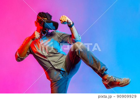 Portrait of concentrated man in shirt virtual reality headset holding clenched fists and raised leg, online gaming. Indoor studio shot isolated on colorful neon light background. 90909929