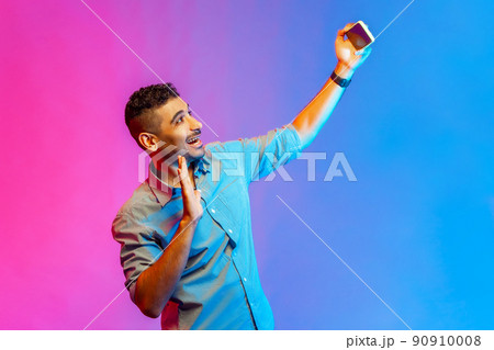 Portrait of friendly man in shirt talking on video call and waving hello gesture, having online conversation on mobile phone. Indoor studio shot isolated on colorful neon light background. Portrait of friendly man in shirt talking on video call and waving hello gesture, having online conversation on mobile phone. Indoor studio shot isolated on colorful neon light background. 90910008