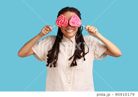 Portrait of playful childish romantic woman with black dreadlocks covering eyes with sugary hear shape candies, wearing white shirt. Indoor studio shot isolated on blue background. 90910179