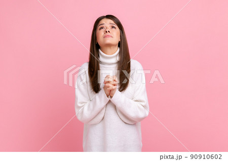Young adult attractive brunette girl looking up with hands in prayer, sincere saying please, apology, wearing white casual style sweater. Indoor studio shot isolated on pink background. Young adult attractive brunette girl looking up with hands in prayer, sincere saying please, apology, wearing white casual style sweater. Indoor studio shot isolated on pink background. 90910602