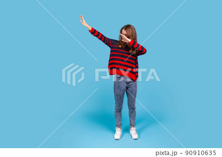 Full length portrait of woman wearing striped casual style sweater, hiding face with dab dance move, performing internet meme of success, dabbing trends. Indoor studio shot isolated on blue background Full length portrait of woman wearing striped casual style sweater, hiding face with dab dance move, performing internet meme of success, dabbing trends. Indoor studio shot isolated on blue background 90910635