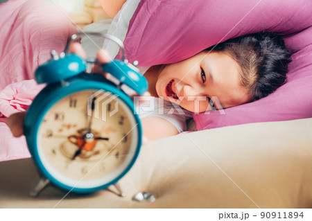 Young beautiful girl hates waking up early in the morning. Sleepy girl looking at alarm clock and trying to hide under the pillow. Students wake up early to go to school. 90911894