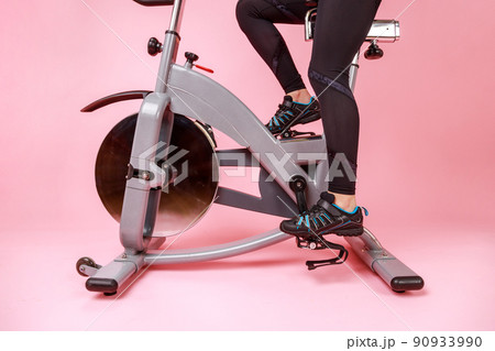 Portrait of the bike simulator and women's legs in black sneakers and leggins, cardio workout, training. Indoor studio shot isolated on pink background. Portrait of the bike simulator and women's legs in black sneakers and leggins, cardio workout, training. Indoor studio shot isolated on pink background. 90933990