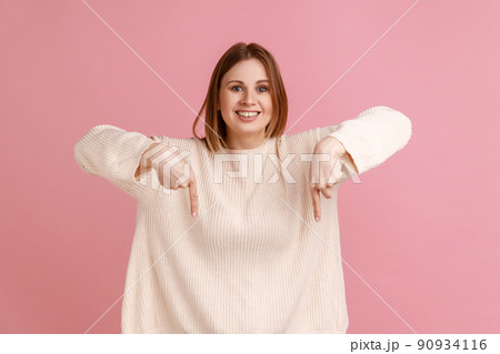 Portrait of attractive happy blond woman pointing down place for commercial idea, looking at camera with toothy smile, wearing white sweater. Indoor studio shot isolated on pink background. Portrait of attractive happy blond woman pointing down place for commercial idea, looking at camera with toothy smile, wearing white sweater. Indoor studio shot isolated on pink background. 90934116