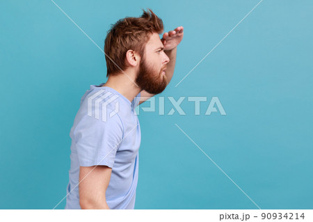 Side view of serious concentrated bearded man looking far away at distance with hand over head, attentively searching for bright future. Indoor studio shot isolated on blue background. Side view of serious concentrated bearded man looking far away at distance with hand over head, attentively searching for bright future. Indoor studio shot isolated on blue background. 90934214