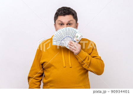 Portrait of dark haired middle aged man covering half of face with many dollar banknotes, looking at camera, wearing urban style hoodie. Indoor studio shot isolated on white background. Portrait of dark haired middle aged man covering half of face with many dollar banknotes, looking at camera, wearing urban style hoodie. Indoor studio shot isolated on white background. 90934401
