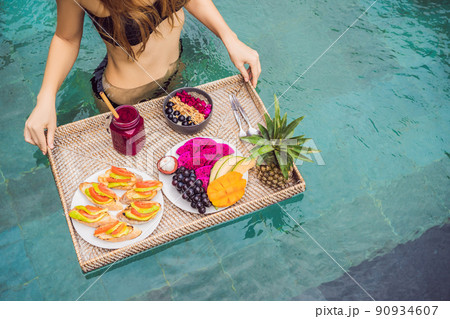 Breakfast tray in swimming pool, floating breakfast in luxury hotel. Girl relaxing in the pool drinking smoothies and eating fruit plate, smoothie bowl by the hotel pool. Exotic summer diet. Tropical 90934607