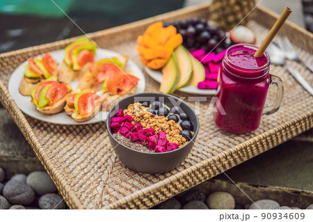 Breakfast on a tray with fruit, buns, avocado sandwiches, smoothie bowl by the pool. Summer healthy diet, vegan breakfast. Tasty vacation concept Breakfast on a tray with fruit, buns, avocado sandwiches, smoothie bowl by the pool. Summer healthy diet, vegan breakfast. Tasty vacation concept 90934609