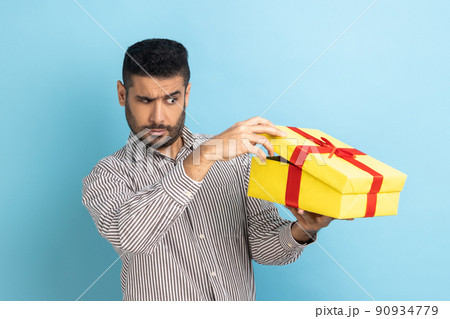 Curious man with beard taking a look inside gift box, unpacking present to know what he received, frustrated with content, wearing striped shirt. Indoor studio shot isolated on blue background. Curious man with beard taking a look inside gift box, unpacking present to know what he received, frustrated with content, wearing striped shirt. Indoor studio shot isolated on blue background. 90934779