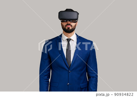 Bearded man standing in vr headset, watching virtual reality movie with calm facial expression, innovative technology, wearing official style suit. Indoor studio shot isolated on gray background. Bearded man standing in vr headset, watching virtual reality movie with calm facial expression, innovative technology, wearing official style suit. Indoor studio shot isolated on gray background. 90934822