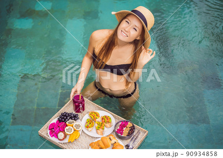 Breakfast tray in swimming pool, floating breakfast in luxury hotel. Girl relaxing in the pool drinking smoothies and eating fruit plate, smoothie bowl by the hotel pool. Exotic summer diet. Tropical 90934858