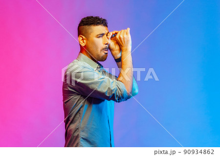 Side view portrait of handsome young adult man in shirt looking far away through monocular gesture, spying shocking content. Indoor studio shot isolated on colorful neon light background. Side view portrait of handsome young adult man in shirt looking far away through monocular gesture, spying shocking content. Indoor studio shot isolated on colorful neon light background. 90934862