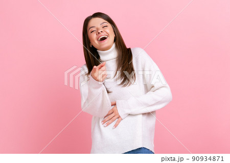 Happy female laughing out loud and pointing finger to camera, hearing funny joke, holding belly, wearing white casual style sweater. Indoor studio shot isolated on pink background. 90934871