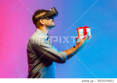 Side view of man in VR virtual reality glasses holding wrapped gift box and smiling joyfully, giving present, celebrating holiday. Indoor studio shot isolated on colorful neon light background. Side view of man in VR virtual reality glasses holding wrapped gift box and smiling joyfully, giving present, celebrating holiday. Indoor studio shot isolated on colorful neon light background. 90934901