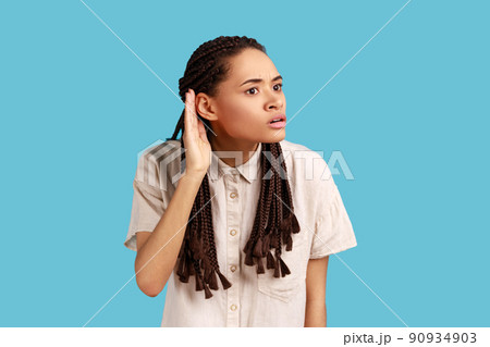 Indignant woman with black dreadlocks tries to overhear someone, holds hand near ear, trying to understand words, eavesdropping keeps mouth opened. Indoor studio shot isolated on blue background. Indignant woman with black dreadlocks tries to overhear someone, holds hand near ear, trying to understand words, eavesdropping keeps mouth opened. Indoor studio shot isolated on blue background. 90934903