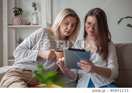 Two young women friends using tablet together, sitting on the couch and smiling Two young women friends using tablet together, sitting on the couch and smiling 90941215
