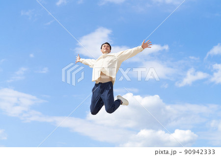 青空のもと両手を広げてジャンプする若い男性 90942333