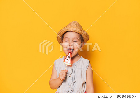 Girl in hat licks watermelon-shaped lollipop against yellow background Girl in hat licks watermelon-shaped lollipop against yellow background 90949907