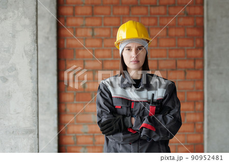 Young serious female engineer in protective helmet and workwear looking at you 90952481