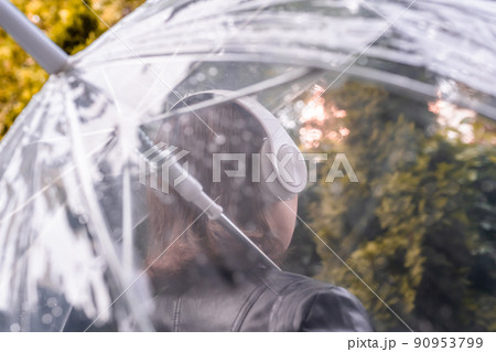 Autumn. Lonely sad woman in a headphones walking in a park, garden. View through wet transparent umbrella with rain drops. Rainy day landscape. Autumn. Lonely sad woman in a headphones walking in a park, garden. View through wet transparent umbrella with rain drops. Rainy day landscape. 90953799