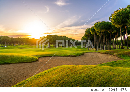 Golf course at sunset with beautiful sky and sand trap. Scenic panoramic view of golf fairway with bunker. Golf field with pines Golf course at sunset with beautiful sky and sand trap. Scenic panoramic view of golf fairway with bunker. Golf field with pines 90954478